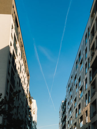 Two airplane contrails crossing in clear blue sky create striking X shape above modern residential buildings, adding dynamic element to urban cityscape. X crossing pattern, geometric, airplane, urbanの写真素材