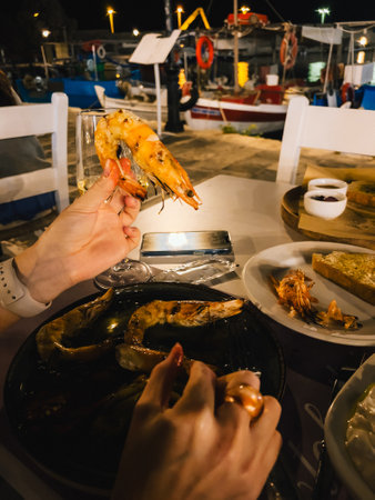POV shot: woman is having dinner at restaurant by the sea, enjoying grilled shrimp and wine under moonlight, with fishing boats in background. Woman dining seaside, savoring grilled shrimp and wineの写真素材