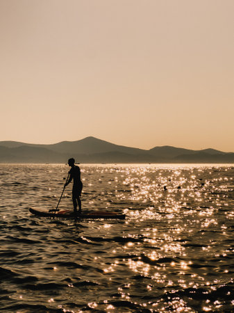 Silhouette of sportsman paddling on stand up paddle board during stunning sunset, with majestic mountain range in background and sunlight reflecting off calm water surface. Paddling, paddle, boardの写真素材