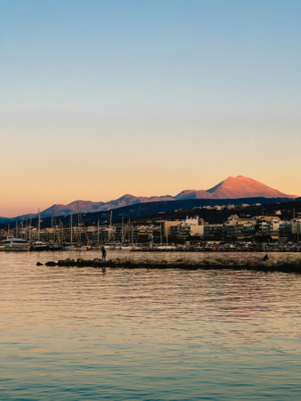 Calm sea reflecting warm hues of sunset sky, featuring Island harbor, breakwater, and majestic...