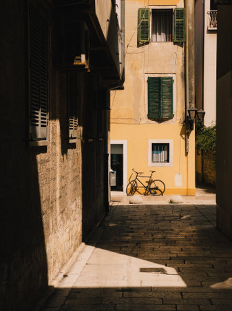 Sunlight illuminating narrow cobblestone alley with yellow wall and black bicycle in Rovinj, Croatia, creating strong shadows on stone pavement. Yellow wall framing vintage bicycle in Rovinj alleywayの写真素材
