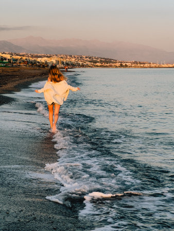Silhouette of woman strolling on shoreline, bathed in soft sunrise glow. Gentle waves, tranquil sea, and fresh breeze enhance peaceful morning walk and feeling of ultimate freedom. cinematic, verticalの写真素材