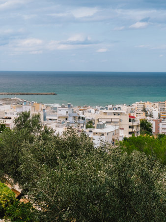 Olive trees frame picturesque view of coastal cityscape nestled against serene Mediterranean Sea on partly cloudy day in Crete, Greece. Olive grove overlooking coastal Cretan cityscape, Mediterraneanの写真素材