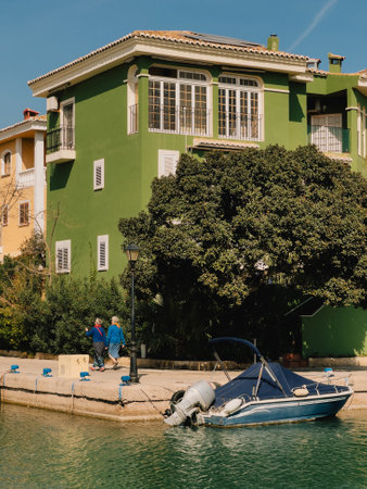 Two senior women walking on paved pathway next to canal with moored boat and green residential building in background on sunny day. Sunlit blue boat moored next to green residential buildingの写真素材
