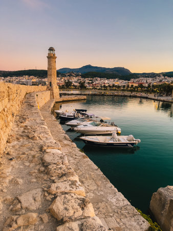 A rugged stone fortress stretches along the coast, framing a picturesque harbor view. Sunset hues, calm water, and city lights add to the nostalgic charm. Weathered stone walls lead toward the seaの写真素材