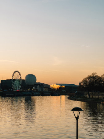 Ferris wheel and balloon lift for sightseeing reflecting on tranquil waters of Vistula River at sunset in Krakow, Poland, with charming street lamp illuminating foreground.の写真素材