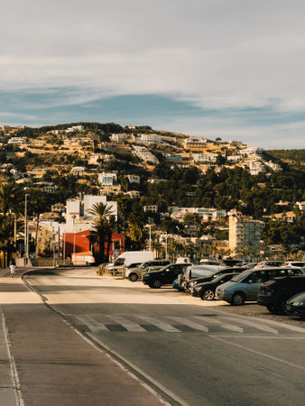 Cars parked along street with buildings covering hill in background with some palm trees and pedestrian crossing in Santa Eularia des Riu, Ibiza, Spain, during sunny day. Parked vehicles, pedestrianの写真素材