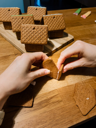 First-person view: Person carefully joining walls of gingerbread house with icing on wooden table, surrounded by other finished houses displayed on cutting board in background. Gingerbread house. POVの写真素材