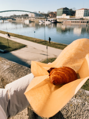 First-person view: tourist is holding delicious croissant just bought in paper bag in sunny day in Krakow, Poland, with Vistula river and modern bridge in background. Person holds crispy croissantの写真素材