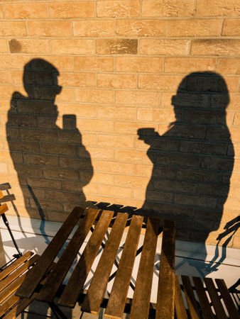 Shadows of two people using smartphones are projected on brick wall behind outdoor wooden table with two chairs, suggesting conversation or social interaction in casual setting. Modern communicationの写真素材
