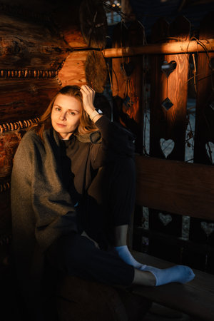 Young woman sitting on wooden porch of traditional log cabin, wrapped in cozy blanket and wearing white short socks, soaking in warm glow of sunset during peaceful evening. Girl enjoying golden warmthの写真素材