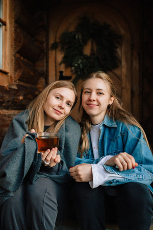 Two young women are sitting on porch of wooden house in countryside, covered with blanket and drinking tea, enjoying quiet moment together. Two rural women relaxing on wooden porch, sharing teaの写真素材