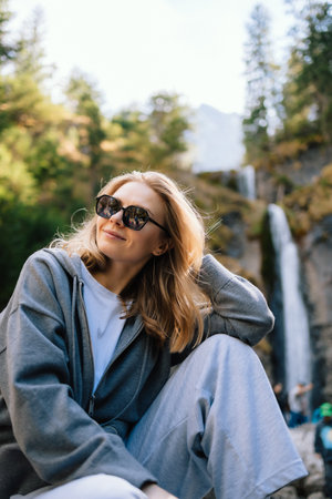 Blonde tourist sitting in front of majestic waterfall, enjoying fresh air and breathtaking mountain views, embodying adventure and relaxation. Blonde woman relaxing near thundering waterfallの写真素材