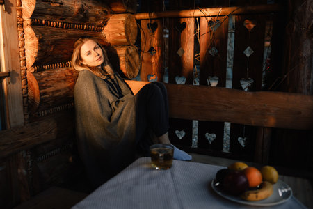 Young woman wrapped in blanket, savoring warm drink and fresh fruits in rustic cottage, as sunset glows through heart shaped cutouts in fence. Woman in cozy atmosphere of country cottage during sunsetの写真素材