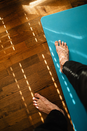 POV shot: man stepping onto turquoise yoga mat on wooden floor, preparing for home workout session in sunlit room, embracing moment of fitness and tranquility. Vertical screen: male practicing yogaの写真素材