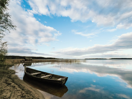 Old wooden fishing boat moored on shore of calm lake reflecting cloudy sky during beautiful sunny day, with small wooden pier and hut on background. Old wooden fishing boat anchored beside serene lakeの写真素材
