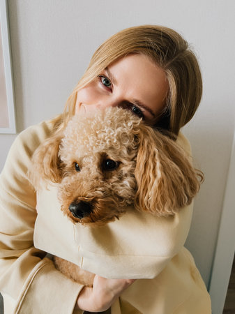 Woman with blond hair, dressed in casual clothes, lovingly embracing her small brown dog, radiating warmth and affection in cozy indoor setting, celebrating their bond. Blond woman hugging brown dogの写真素材