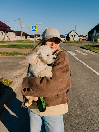 Casually dressed young woman enjoying sunny day while holding her small curly white dog on suburban...