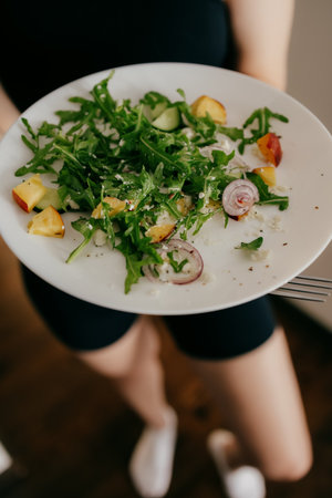 Woman is holding white plate with fresh arugula salad, peach slices, cucumber, red onion, and crumbled cheese, promoting healthy and delicious meal. Woman holding white plate with green healthy saladの写真素材