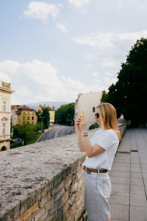 Young blonde woman tourist wearing white shirt and sunglasses is taking pictures of cityscape using her smartphone, enjoying summer vacation. Blonde traveler photographing urban landscape with phoneの写真素材