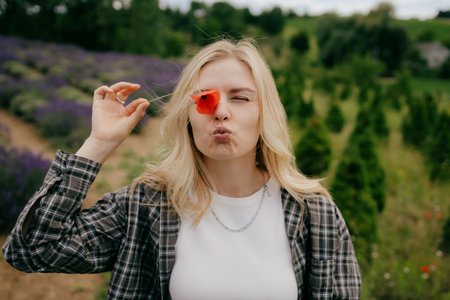 Young blonde female farmer is holding red poppy flower in front of her eye and pouting in lavender field, enjoying moment of playful joy amidst serene landscape. Farmer covering eye with red poppyの写真素材