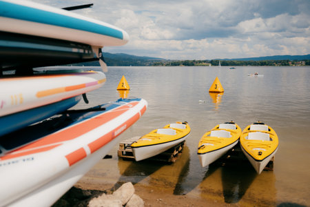 Yellow kayaks and colorful paddle boards are stored on shore of lake on partly cloudy summer day, while canoeist practices in distance. Colorful watercraft resting on lakeside shore next to sap boardsの写真素材