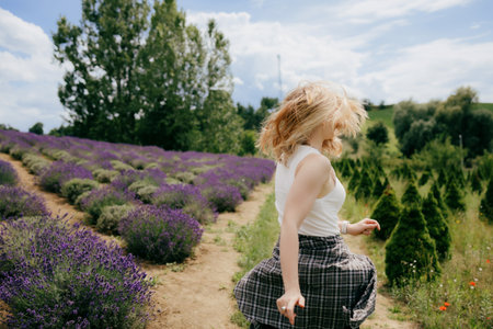 Blonde woman wearing white tank top and plaid shirt is twirling and running through rows of lavender on sunny day, expressing joy and happiness. Joyful blonde woman dancing amid purple lavender fieldsの写真素材