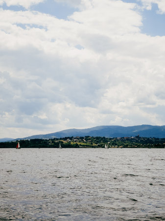 Sailboats are peacefully navigating lake on partly cloudy day, with mountains and small village visible in background, creating serene and picturesque scene. Sailboats gliding across calm lake watersの写真素材