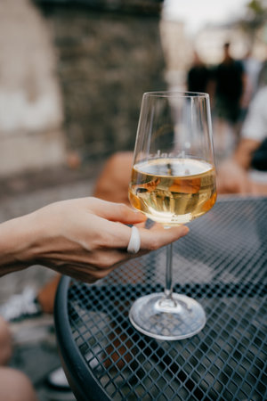 Woman with white ring holding glass of chilled white wine, sitting at metal table in an outdoor cafe, savoring tranquil moment under warm sunlight. Woman wearing white ring grasping white wine glassの写真素材