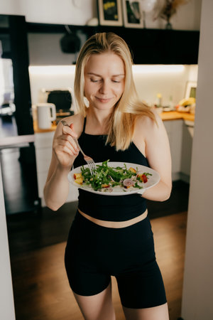 Blond woman wearing sportswear is holding plate of fresh mixed salad and eating it with closed eyes and delightful expression in her modern kitchen. Sporty woman enjoying mixed salad. Healthy lifeの写真素材