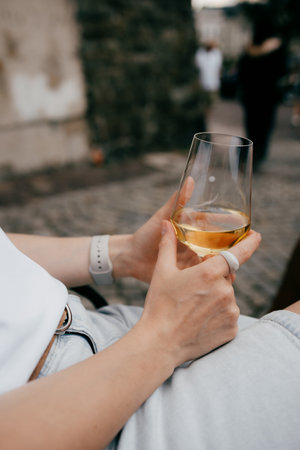 Woman sitting outdoors in casual attire, savoring glass of chilled white wine while enjoying peaceful moment of relaxation amidst vibrant urban backdrop. Woman relaxing outdoors, holding white wineの写真素材