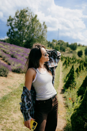 Woman relaxing in lavender field, wearing sunglasses, casual attire, capturing scenic landscape with smartphone while enjoying warm sunlight and fragrant surroundings. Woman posing in lavender fieldの写真素材