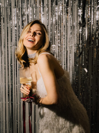 Young blonde woman wearing white dress and necklace is laughing and holding glass of white wine in...