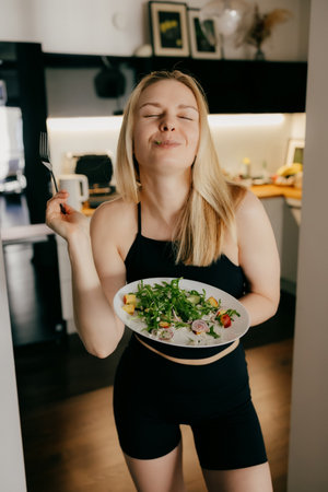 Fit young woman savors healthy salad in her modern kitchen, eyes closed in pure enjoyment as she takes bite, embracing nutritious and delicious meal. Fit female enjoying vibrant green saladの写真素材