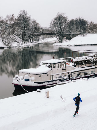 Man is jogging along snow covered riverbank, passing moored boat blanketed in snow, with bridge and snow laden trees in background during serene winter day. Male runs traversing snow covered riverbankの写真素材