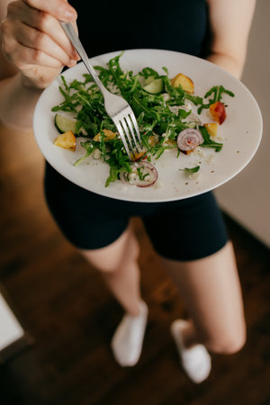 Woman wearing sportswear holding plate with fork and eating fresh healthy salad with arugula, cucumber, peaches and feta cheese, healthy lifestyle and eating habits concept. Wellness focused lifestyleの写真素材