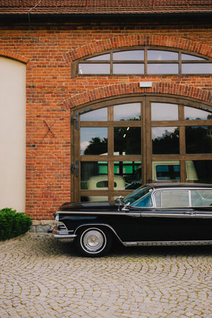 Shiny black limousine parked on cobblestone driveway in front of vintage brick building with large wooden doors, showcasing luxury and timeless style. Black luxury limousine beside vintage brick houseの写真素材
