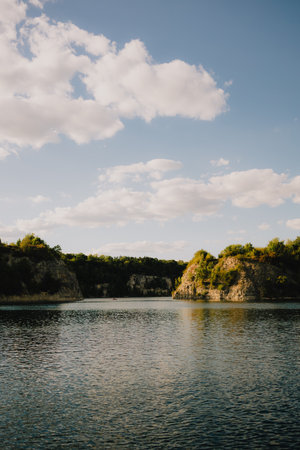 Calm lake water reflecting bright sky filled with fluffy white clouds, surrounded by rocky hills...