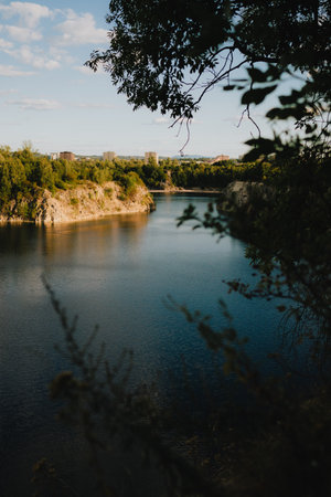 Sun shining on calm quarry lake reflecting cloudy sky, framed by lush green foliage and trees, creating serene and peaceful atmosphere. Quarry water, mirroring cloudy skies amid verdant greeneryの写真素材