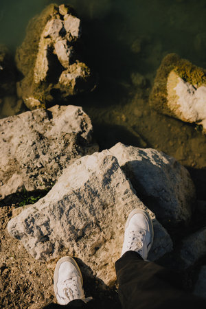First-person view: person standing on large rocks by tranquil body of water, wearing white sneakers...