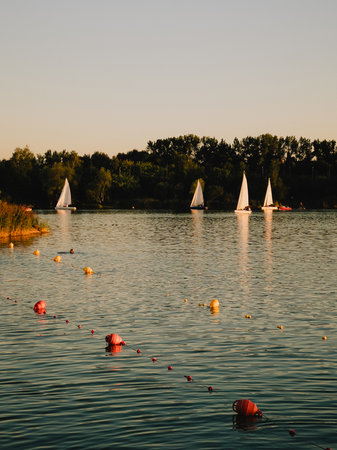 Sailboats are sailing on calm lake at sunset, with line of buoys in foreground marking swimming...