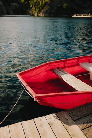 Red rowboat with wooden seats gently rocking on calm lake waters, secured to wooden dock, creating...