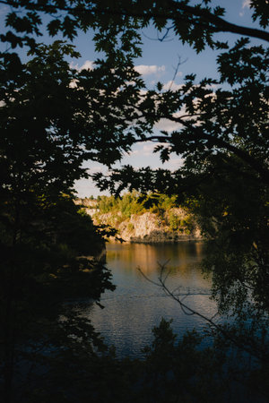 Warm hues of setting sun reflect on calm surface of lake nestled amidst rocky landscape, creating serene scene viewed through natural frame of verdant leaves. Tranquil lake surface, rocky terrainの写真素材