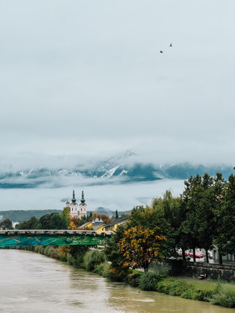 Scenic view of Salzach river flowing through Salzburg city center with colorful bridge, church towers and snow capped Alps mountains rising above low clouds in the background on cloudy autumn dayの写真素材