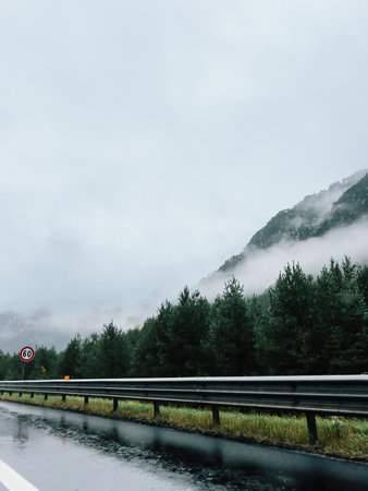 Wet highway winding through mountain forest, enveloped in fog on rainy day, featuring speed limit...