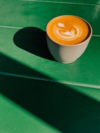 Ceramic cup filled with creamy cappuccino, featuring intricate latte art foam, resting on vibrant green table in cozy cafe, casting striking shadows from bright sunlight. Ceramic cappuccino cupの写真素材