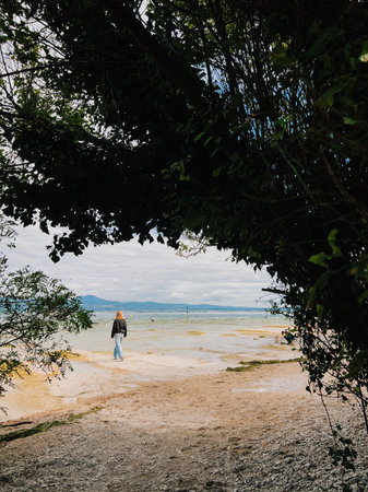 Blonde woman walking on shore of lake framed by branches and leaves of trees and bushes, enjoying...