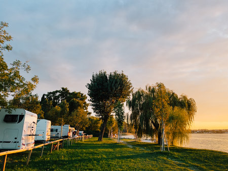 Campers parked by serene lake, soaking in warm glow of golden hour sunset, crafting tranquil and picturesque vacation scene filled with joy and relaxation. Camping trailer beside calm lake watersの写真素材