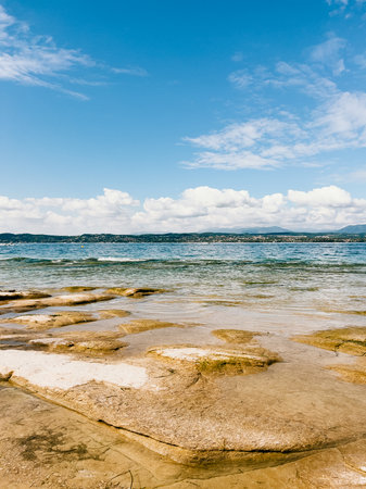 Flat rocks reflecting sunlight on shores of Lake Garda, showcasing beautiful summer day. Calm waters and blue sky dotted with fluffy clouds create tranquil, scenic atmosphere. Lake Garda shorelineの写真素材