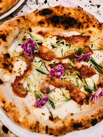 Round gourmet pizza topped with zucchini flowers, crispy bacon, and rich creamy sauce resting on...